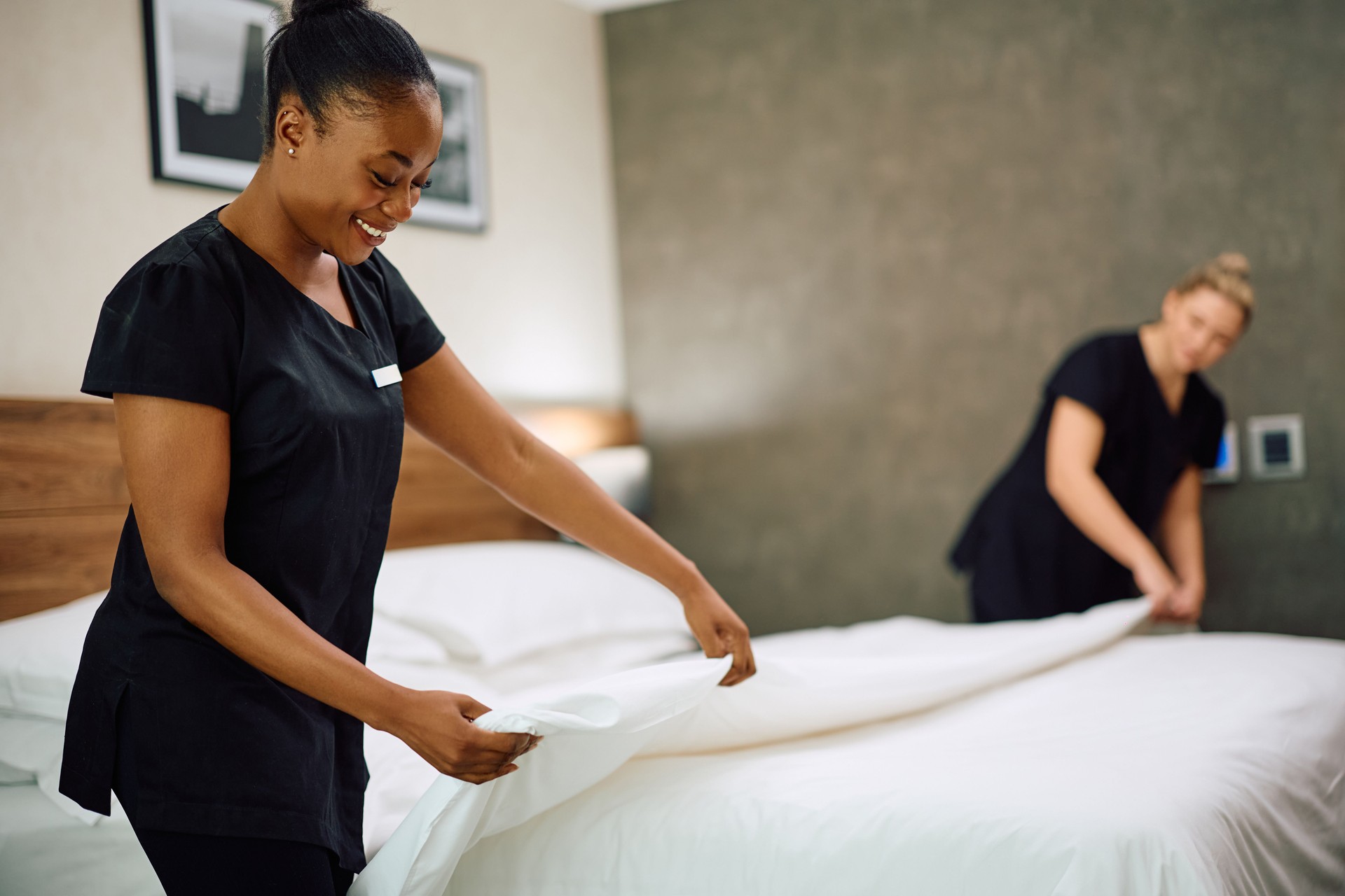 Happy black housekeeper and her female colleague making bed in hotel room.